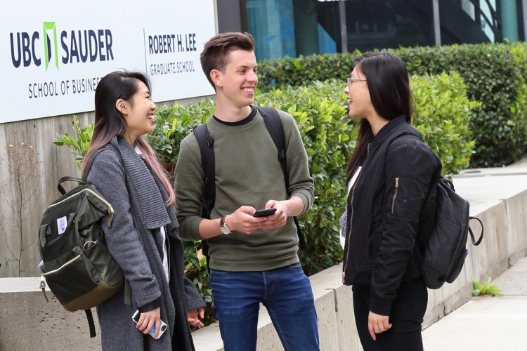 Three students wearing backpacks laugh together in front of the UBC Sauder School of Business sign